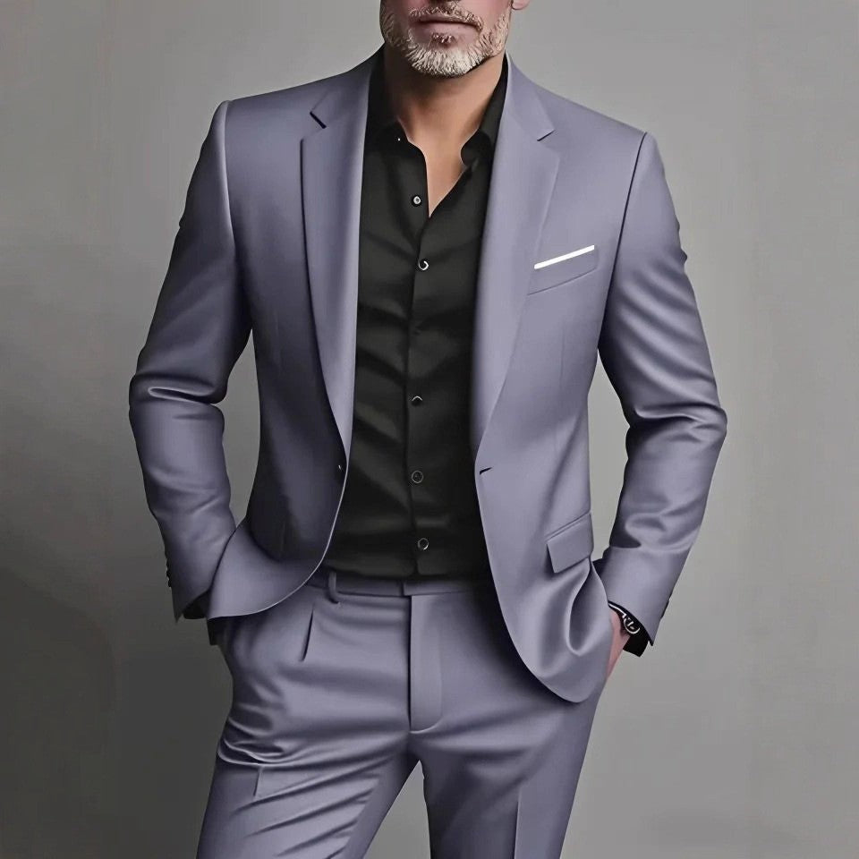 A man wearing a slim-fit light purple-gray suit with a black button-down shirt, standing against a neutral gray background.