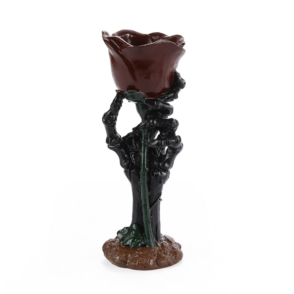 A gothic skeleton hand holding a dark red rose stem, sculpted as a decorative candle holder or vase, set against a plain white background.