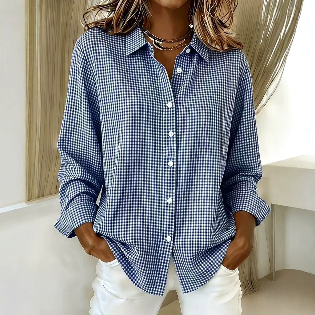 A woman wearing a blue and white gingham check long-sleeve button-up shirt with white pants, standing indoors near a window with light-colored curtains.