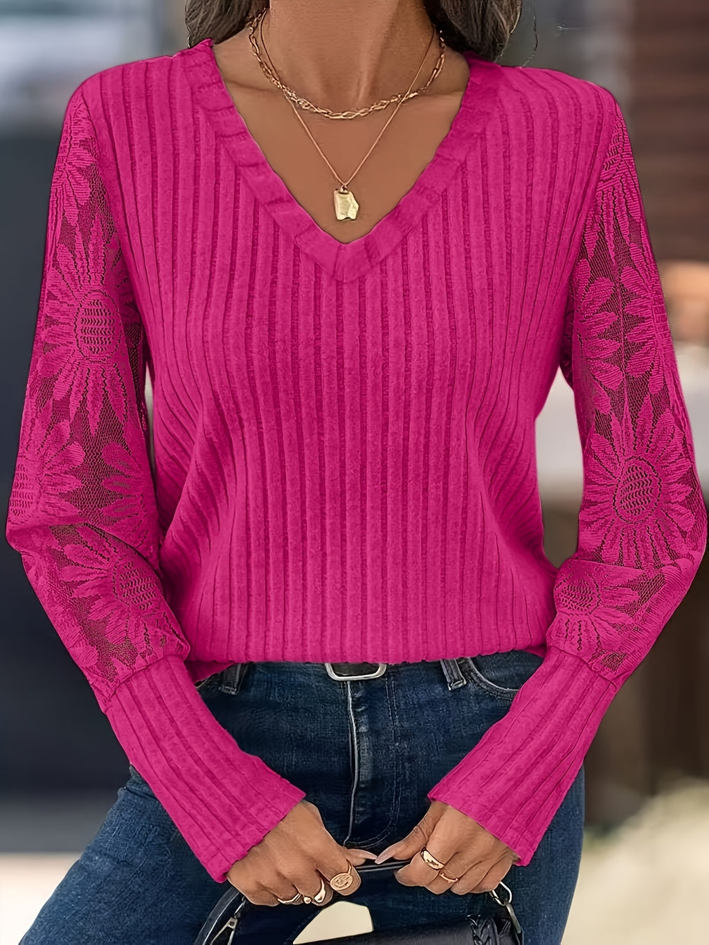 A woman wearing a bright pink ribbed knit sweater with floral lace sleeves, paired with blue jeans and layered gold necklaces.