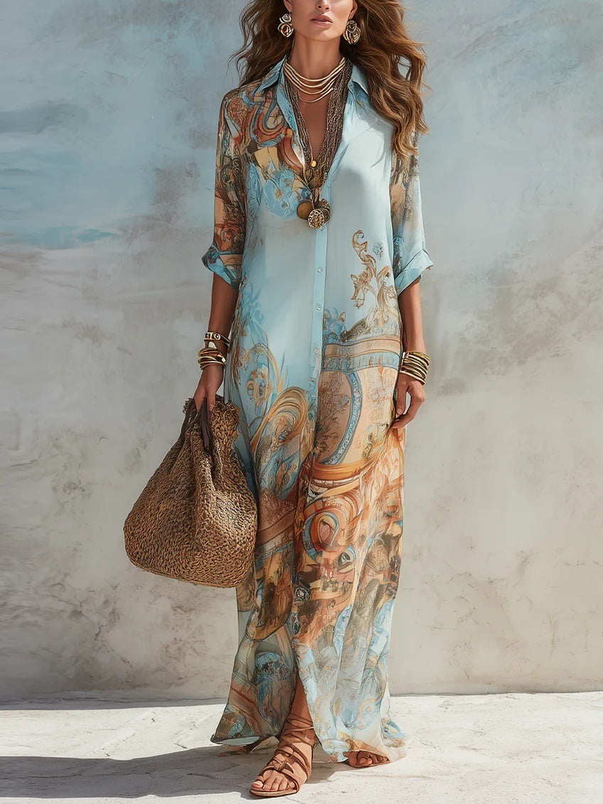 A woman models a light blue maxi shirt dress with a detailed classical art print in brown, gold, and beige. She is accessorized with layered gold necklaces, bracelets, and holds a brown woven tote bag against a textured beige and blue wall.