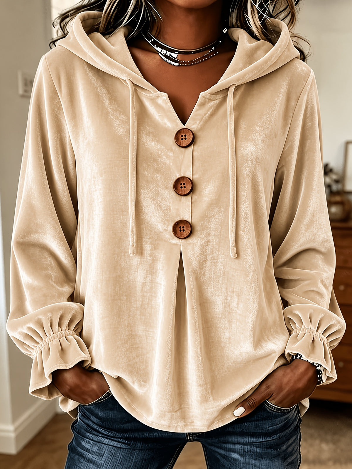 A woman wearing a beige velvet hooded pullover top with three large brown buttons, drawstrings, and ruffled cuffs, paired with dark blue jeans.