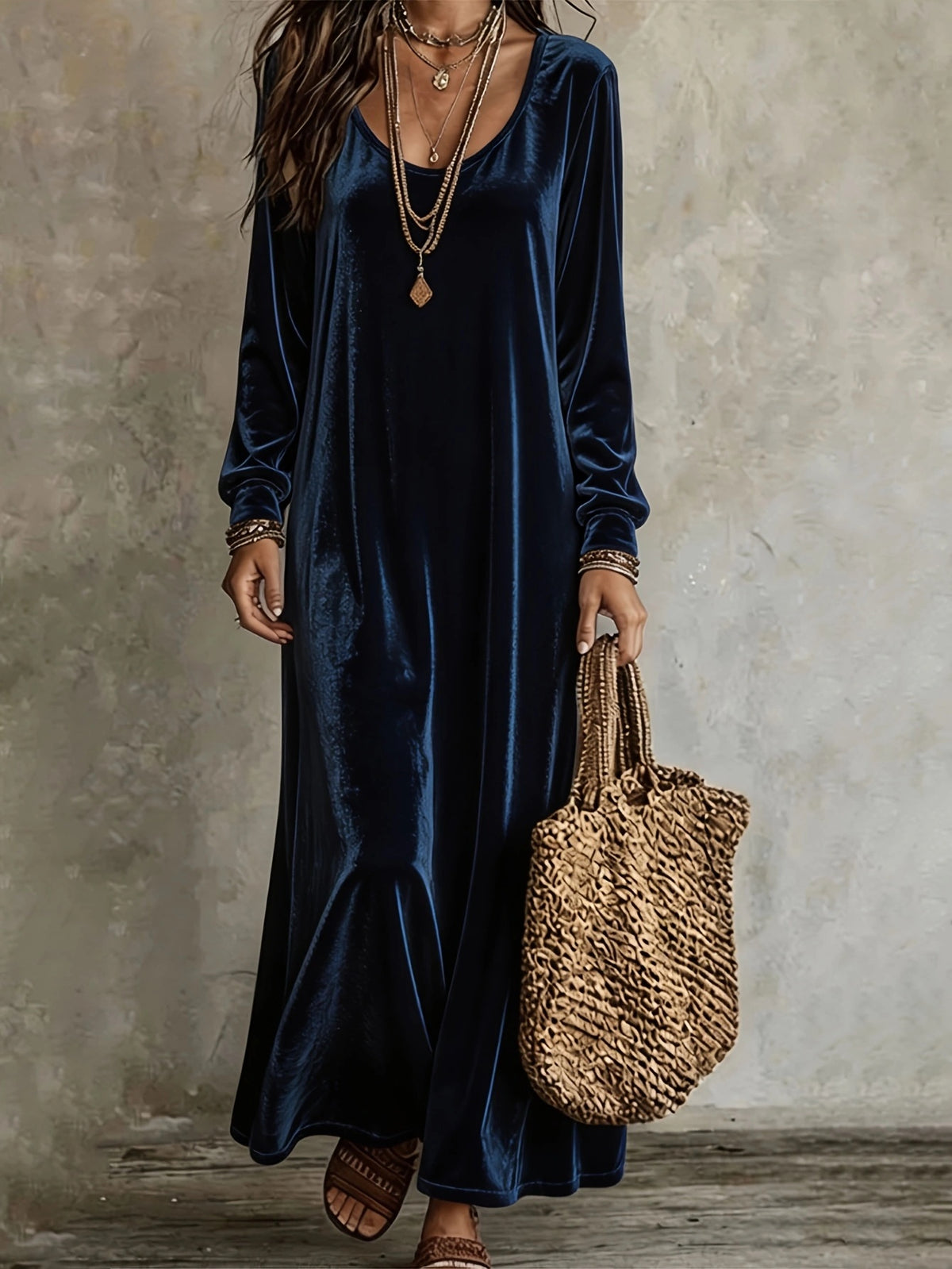 A woman wearing a long-sleeved navy blue velvet maxi dress, accessorized with layered gold necklaces, bracelets, and a woven straw tote bag, standing against a textured gray concrete wall.