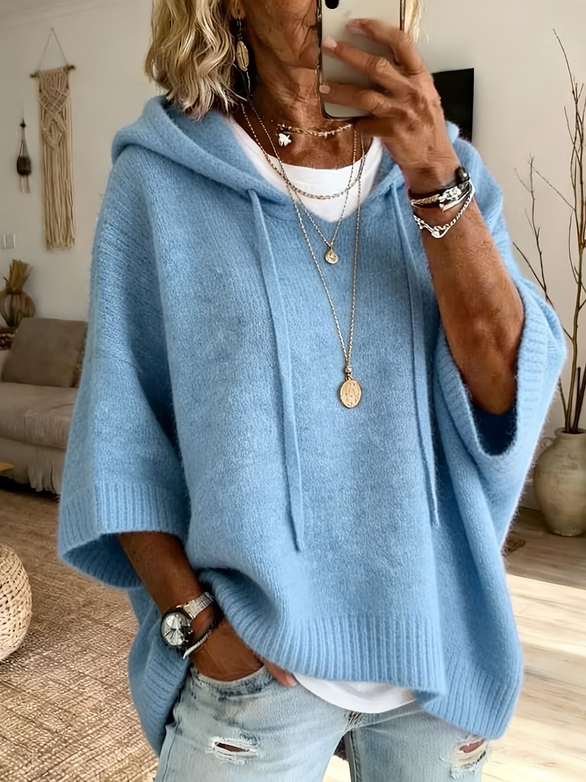 A woman takes a mirror selfie wearing a light blue knit poncho hoodie with drawstrings, layered over a white t-shirt and light-wash distressed jeans. She is accessorized with layered gold necklaces, bracelets, and a watch.