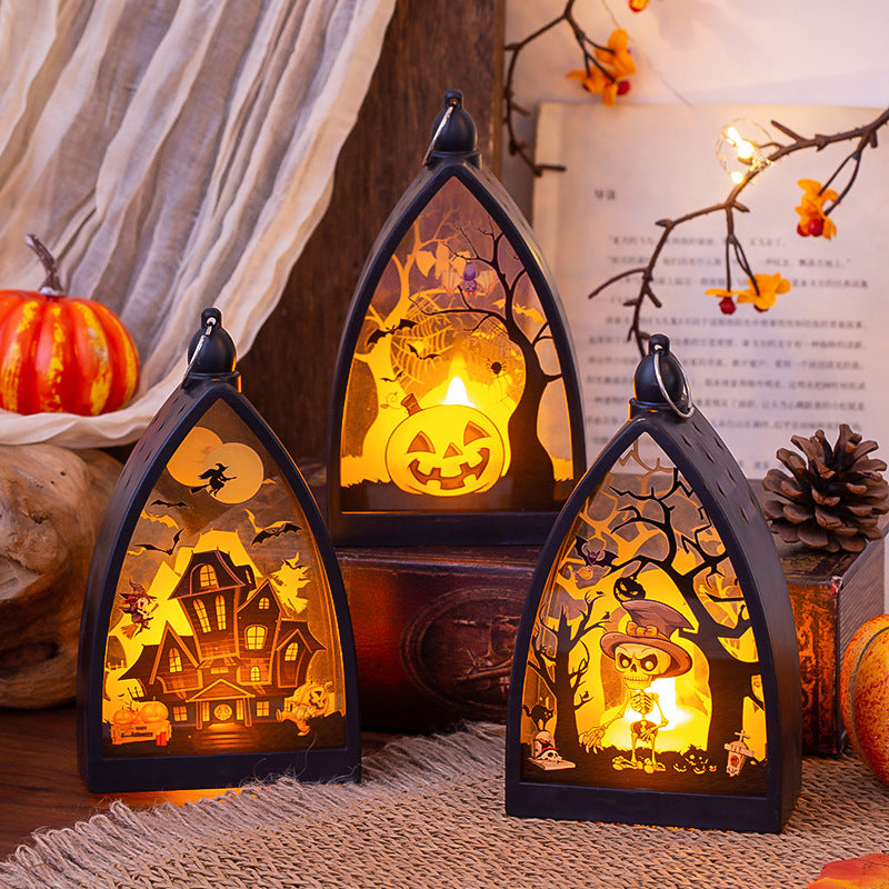 Three gothic-style Halloween lanterns with black arched frames, lit from within to show spooky cutout scenes including a jack-o'-lantern, haunted house, and owl under a tree, arranged on a rustic tabletop with pumpkins, burlap, and autumn decor in the background.