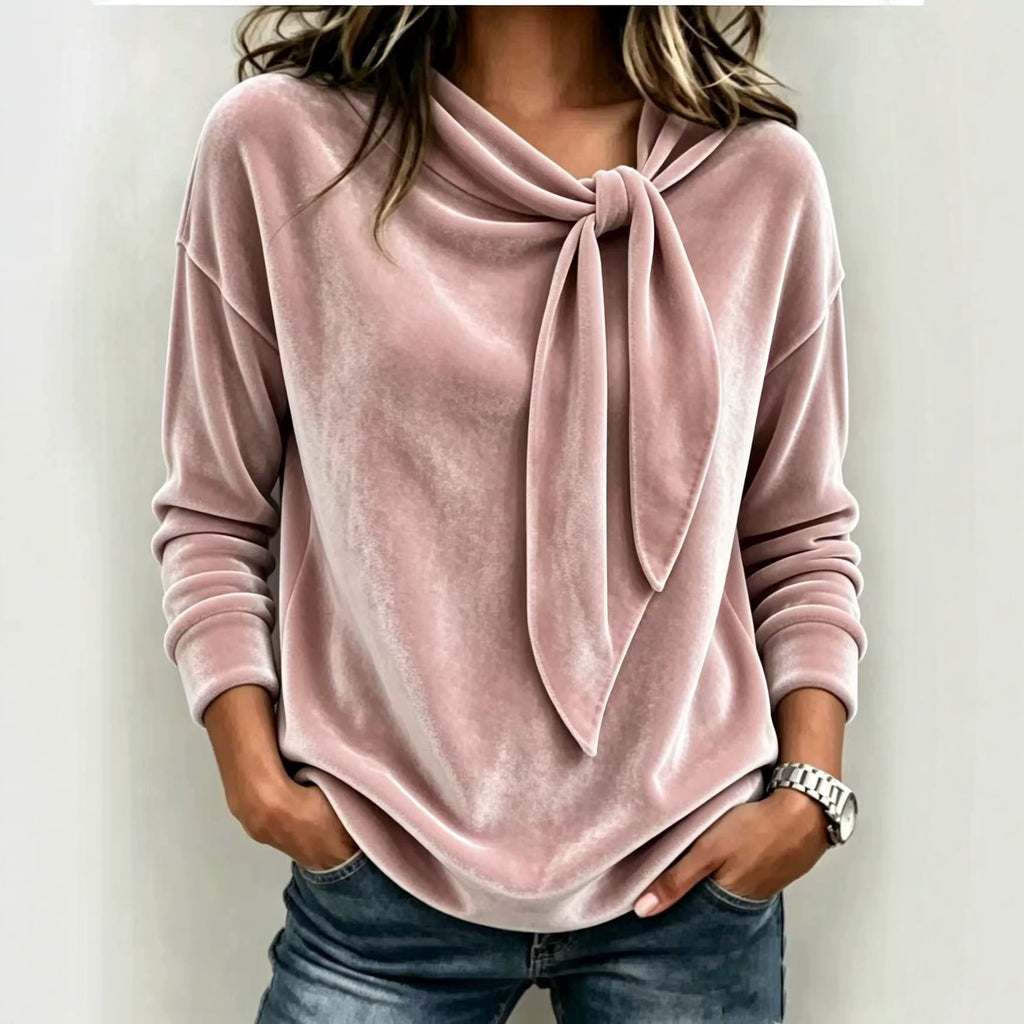 A woman wearing a dusty pink velvet long-sleeve top with a knotted tie neckline, paired with blue jeans, shown against a plain light background.