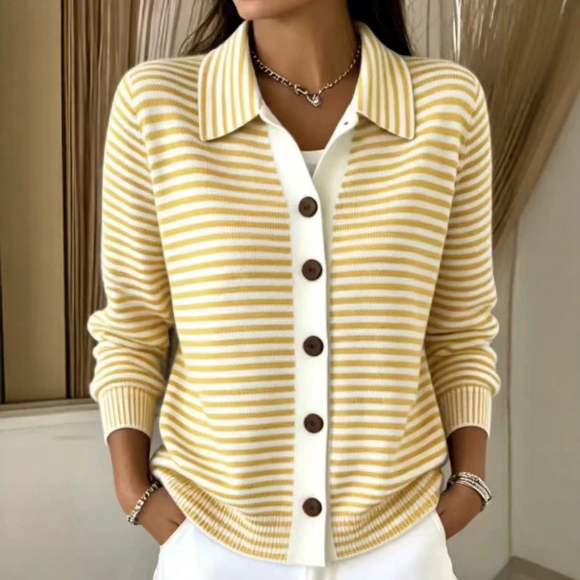 A woman wearing a yellow and white striped knit cardigan with a white placket and dark brown buttons, paired with white pants and jewelry, in an indoor setting.