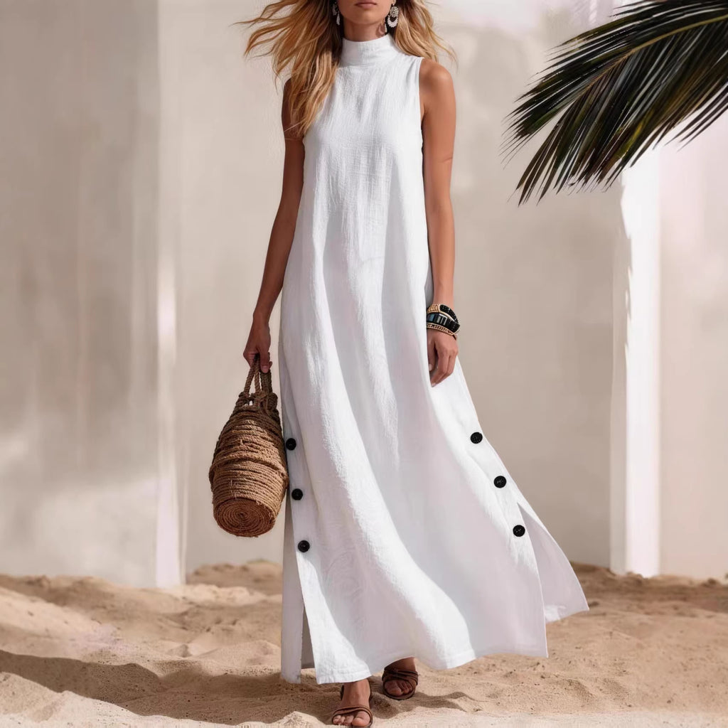 Woman in a white sleeveless dress with black buttons standing on sand with palm leaves.