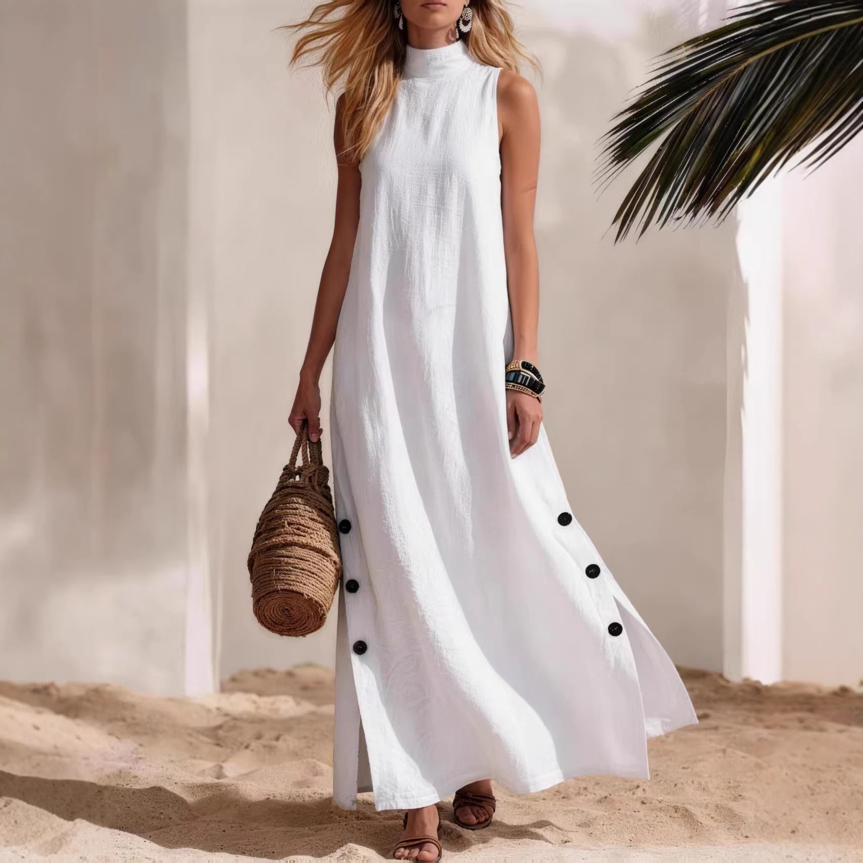 Woman in a white sleeveless dress with black buttons standing on sand with palm leaves.