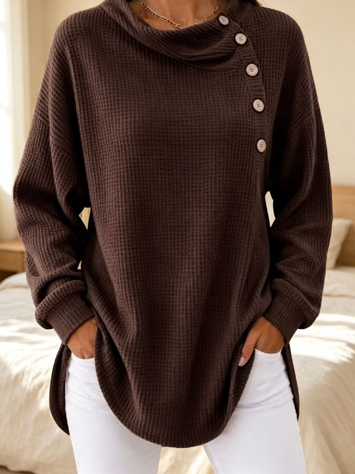 A woman wearing a dark brown waffle-knit cowl neck sweater with an asymmetrical button placket, paired with white pants, standing in a bedroom setting.