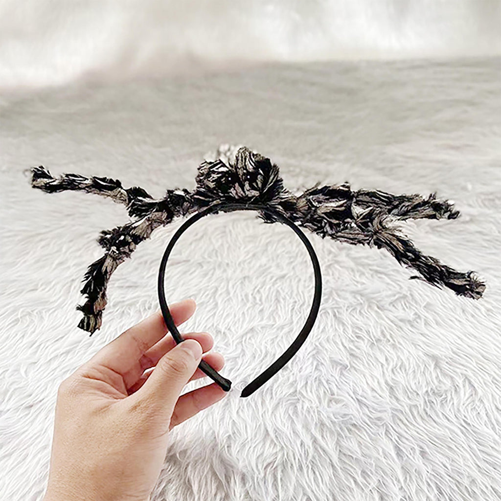 A hand holds a black headband decorated with a large, fluffy black and gray feather-like bow against a white faux fur background.
