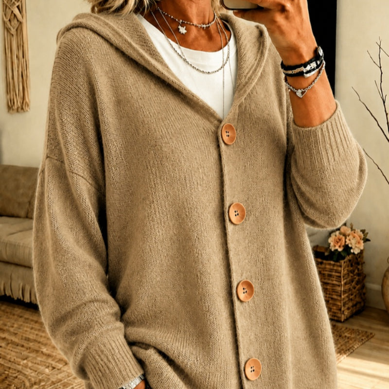 A person wearing a cozy oversized knit cardigan with a hood and large brown buttons, layered over a white top in a warm indoor setting.