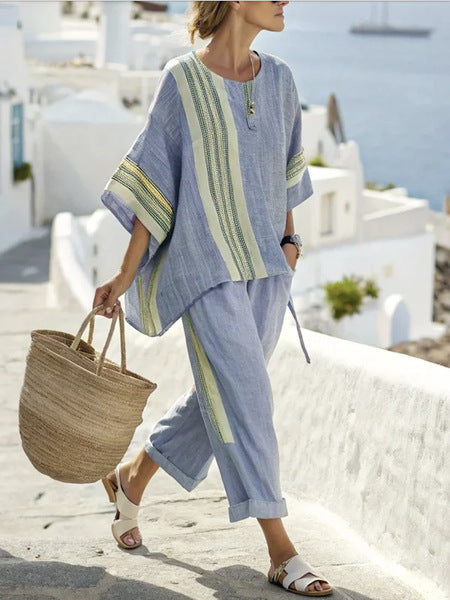 Woman in a blue and green outfit holding a straw bag on a sunlit street with white buildings.