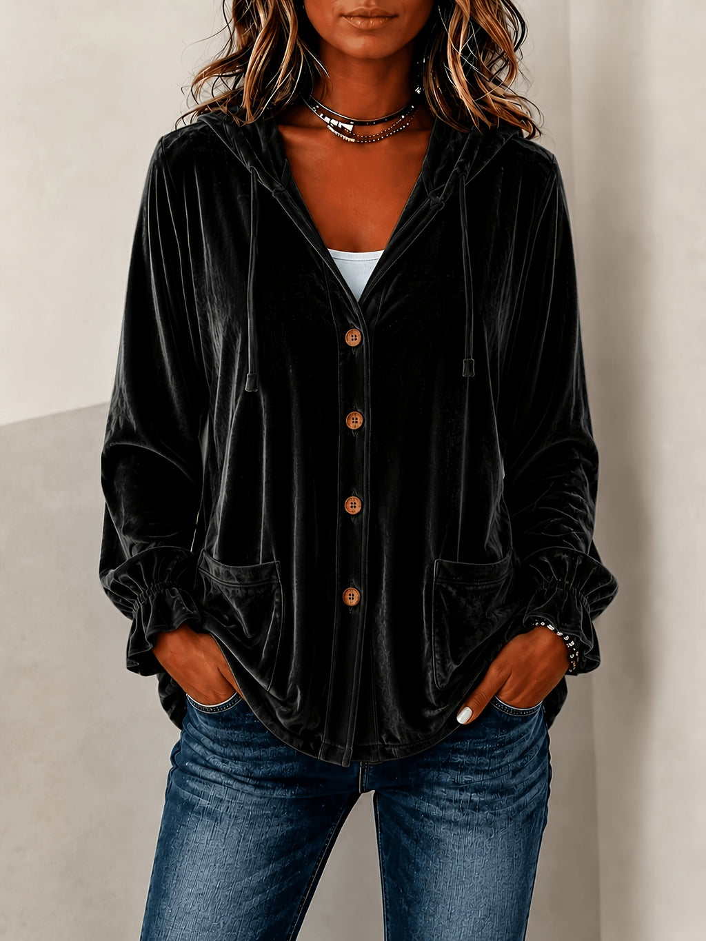 A woman wearing a black velvet button-up hooded jacket with brown buttons, paired with blue jeans, shown against a neutral light gray background.