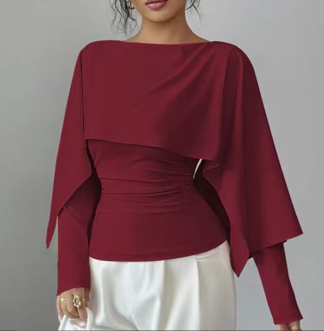 A woman wearing a deep burgundy long-sleeve blouse with a draped, asymmetrical layered overlay and gathered waist detail, paired with white pants, against a plain gray background.