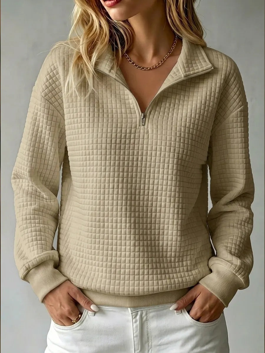 A woman wearing a beige quilted quarter-zip pullover sweater with a stand collar, paired with white pants, shown against a neutral gray background.