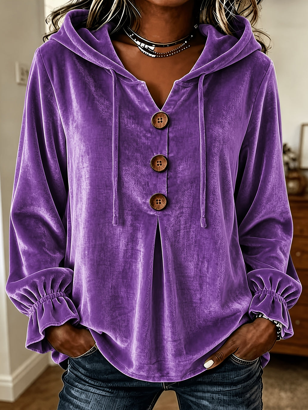 A woman wearing a purple velvet hooded long-sleeve top with three large brown buttons down the front, elasticized ruffled cuffs, and a split neckline, paired with dark blue jeans.