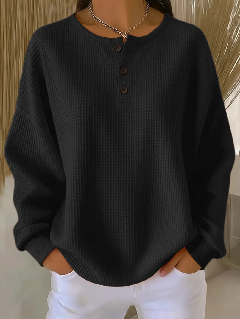 A person wearing a dark charcoal waffle-knit long-sleeve henley top with three dark buttons, paired with white pants, shown from the neck down in an indoor setting.
