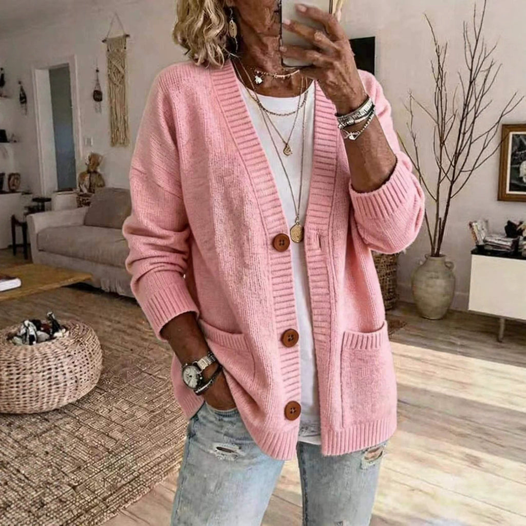 A woman takes a mirror selfie in a cozy living room, wearing a pink knit cardigan with brown buttons over a white top and light blue jeans.