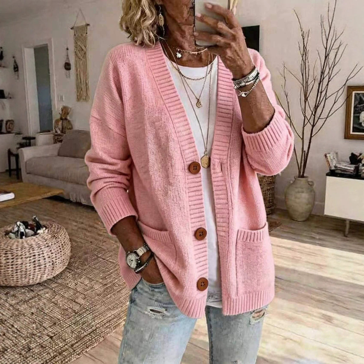 A woman takes a mirror selfie in a cozy living room, wearing a pink knit cardigan with brown buttons over a white top and light blue jeans.