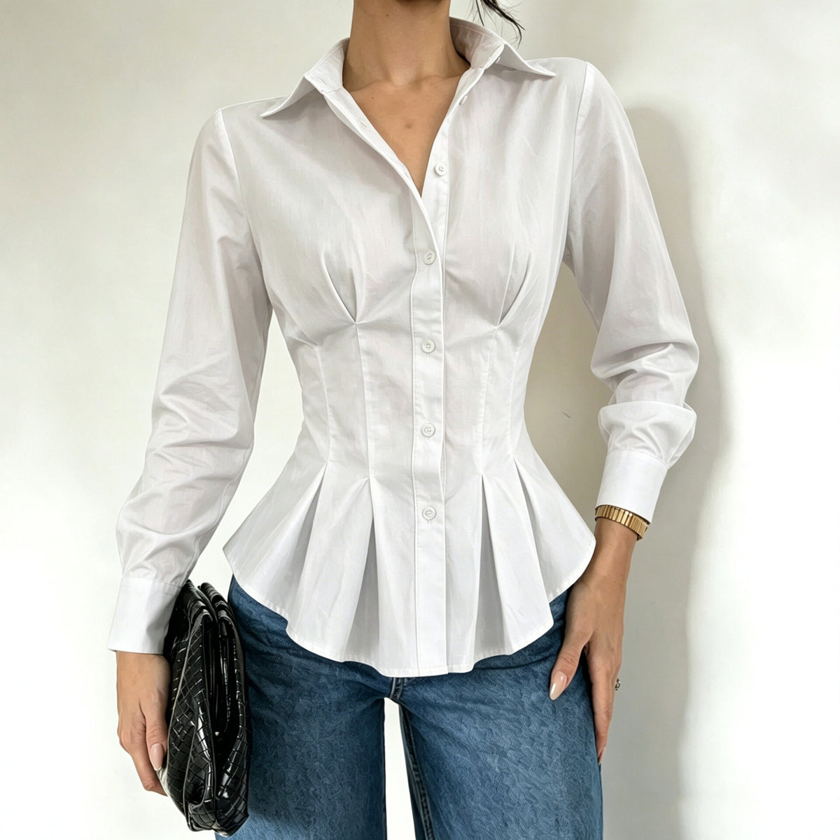 A woman wearing a fitted white long-sleeve button-up blouse with a peplum hem and structured shoulders, paired with blue jeans and holding a black clutch against a plain light background.