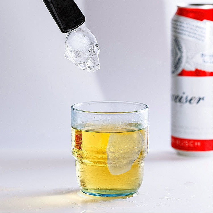 Glass of yellow liquid with a ice cube being added, next to a can of beer on a light gray background