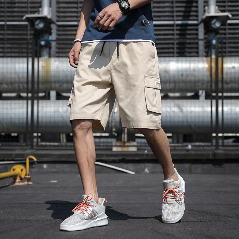 Person wearing beige cargo shorts and white sneakers with orange laces, standing on a dark surface with industrial background.