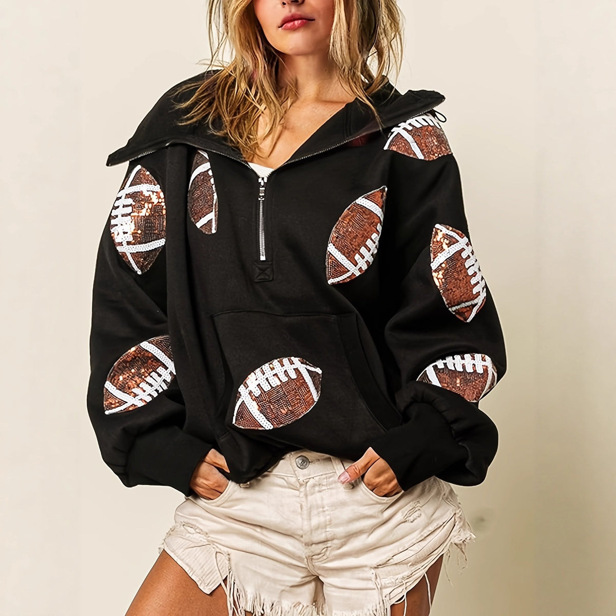 A woman wearing a black pullover hoodie with brown and white football print patches on the sleeves and front pocket, paired with light beige shorts, against a plain beige background.