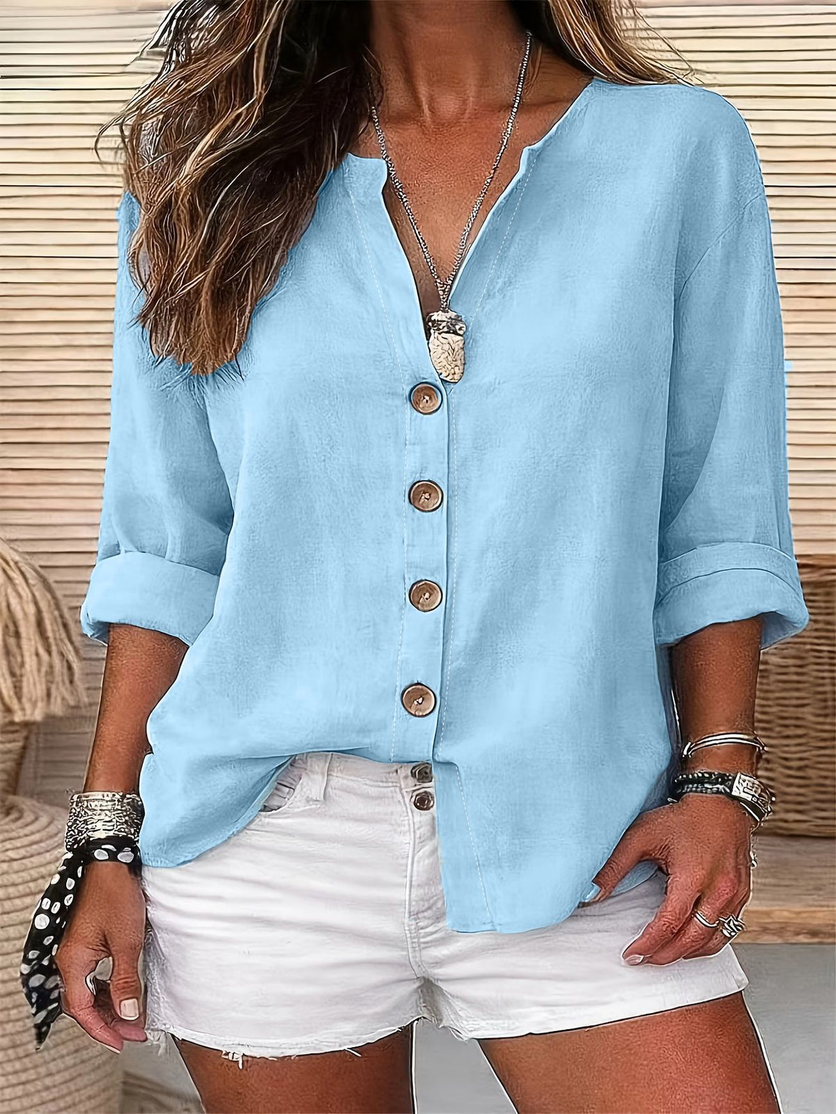 A woman wearing a light blue linen button-up blouse with rolled sleeves and white denim shorts, accessorized with a silver pendant necklace and bracelets.