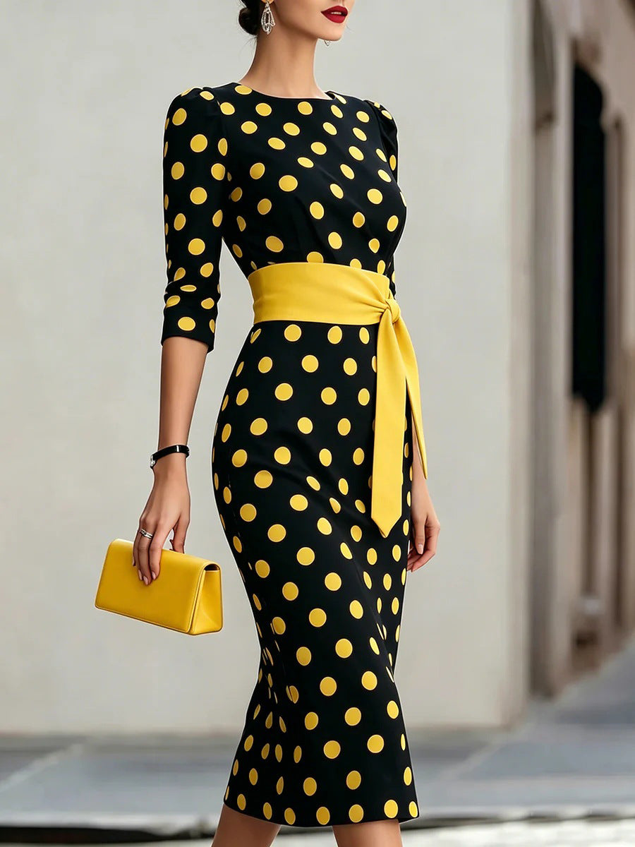 Woman wearing a black dress with yellow polka dots and a matching yellow belt, holding a yellow clutch.