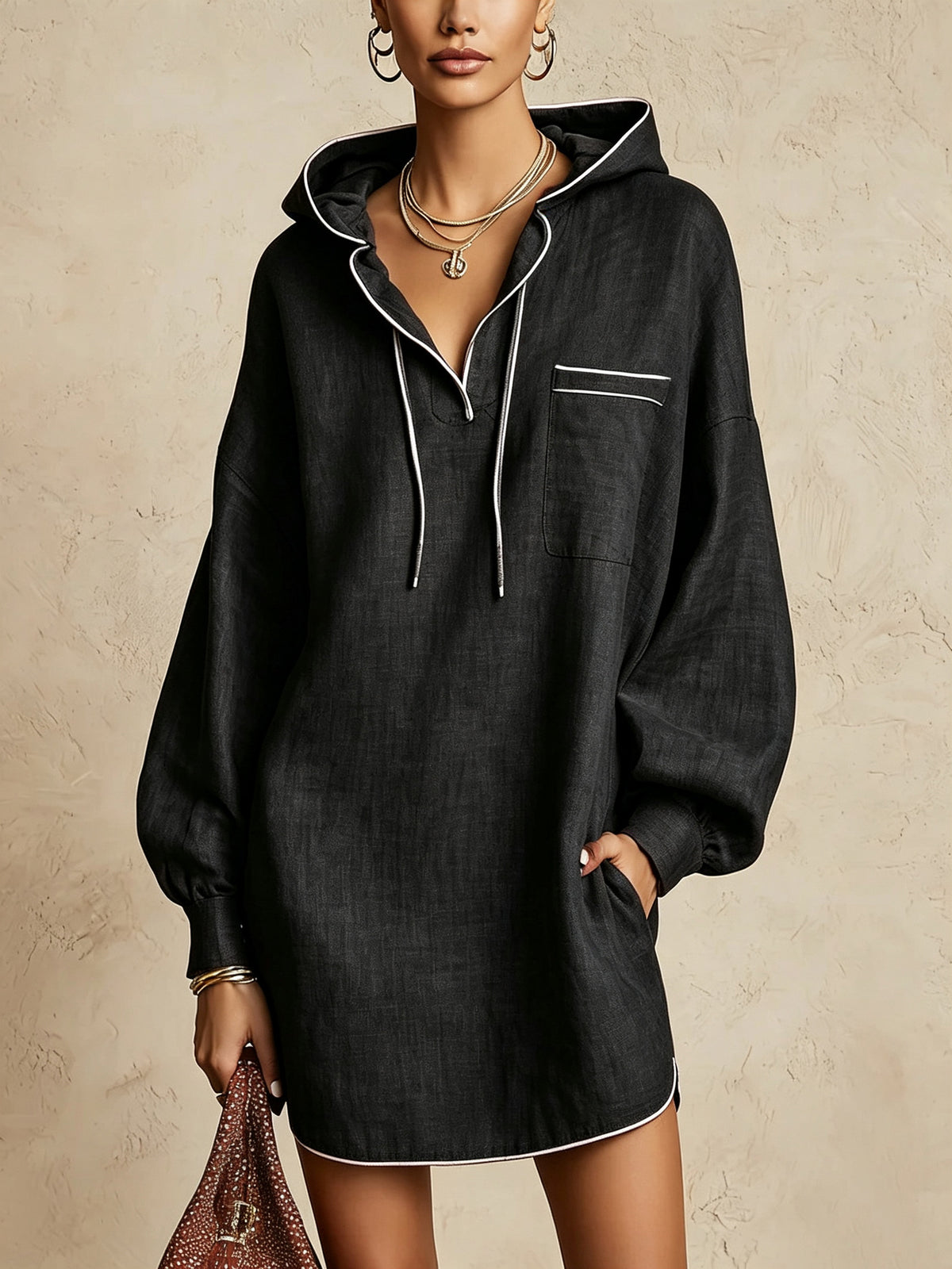 A woman models an oversized black hooded shirt dress with white contrast piping and drawstrings, set against a textured beige wall.