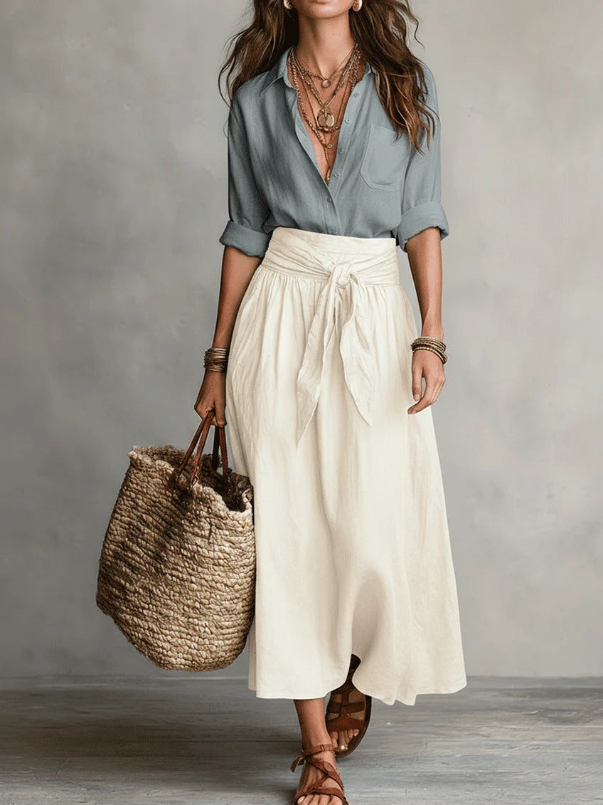 Woman wearing a light blue shirt and white skirt, holding a woven bag against a neutral background