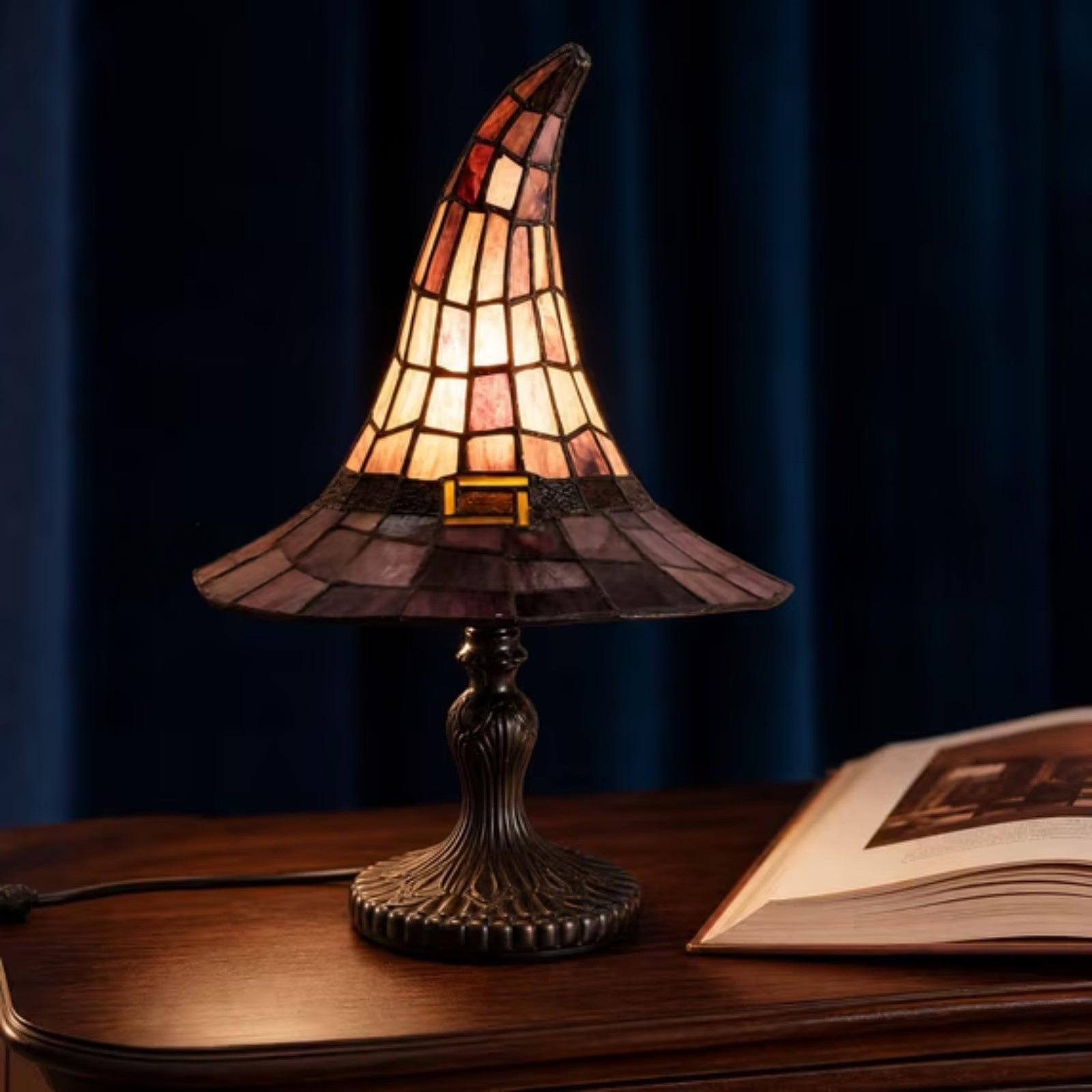 A stained-glass-style table lamp shaped like a witch hat sits on a wooden table against a dark blue curtain background. An open book is partially visible on the right, and the lamp is turned on, casting a warm glow.