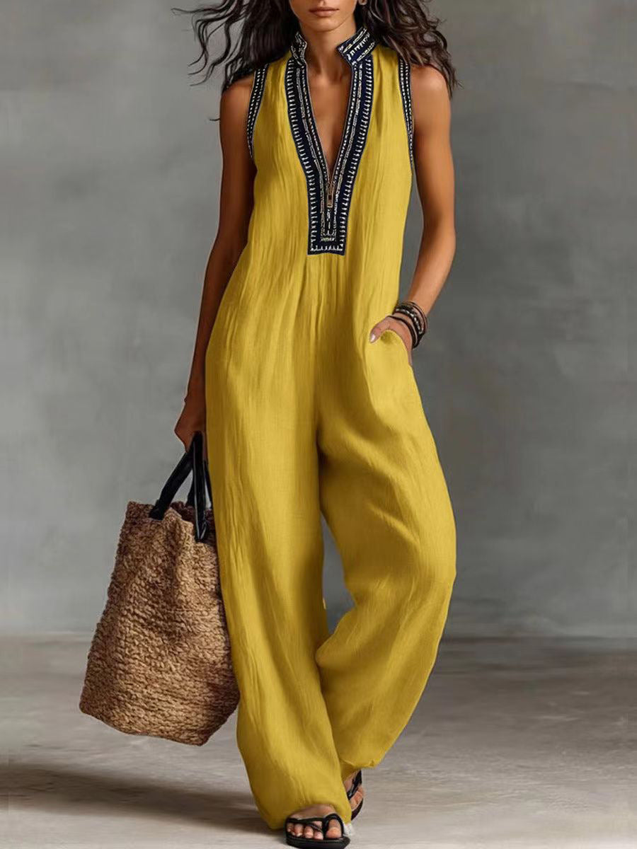 Woman wearing a yellow jumpsuit with black embroidery, holding a woven bag against a neutral background.