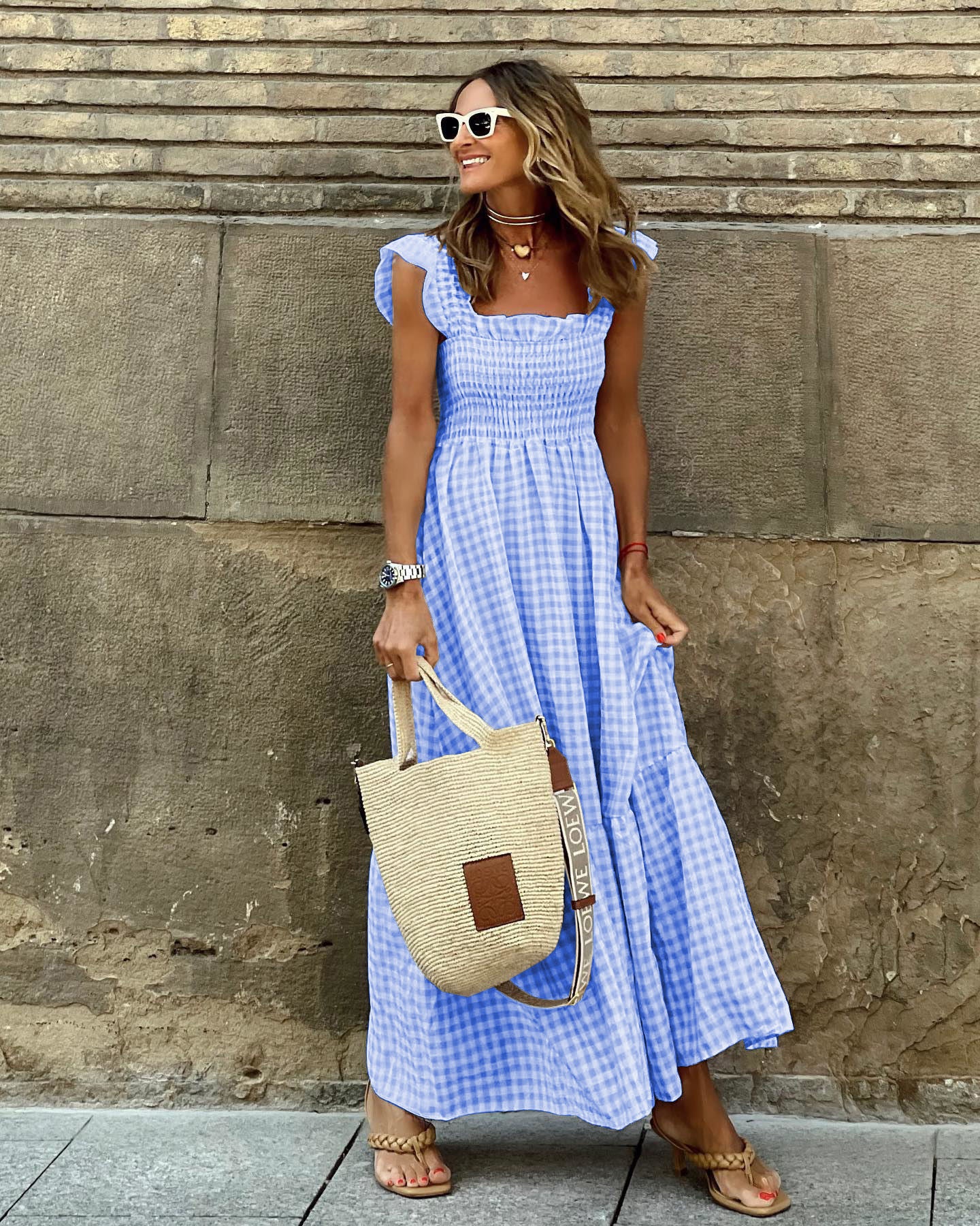 Woman in a blue checkered dress holding a straw bag against a stone wall.