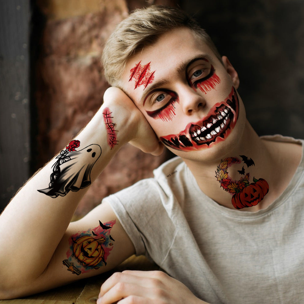 A young man with Halloween face makeup featuring a creepy grin and red scratch marks, wearing temporary tattoos on his arms and neck, resting his head on his hand against a dark rustic background.