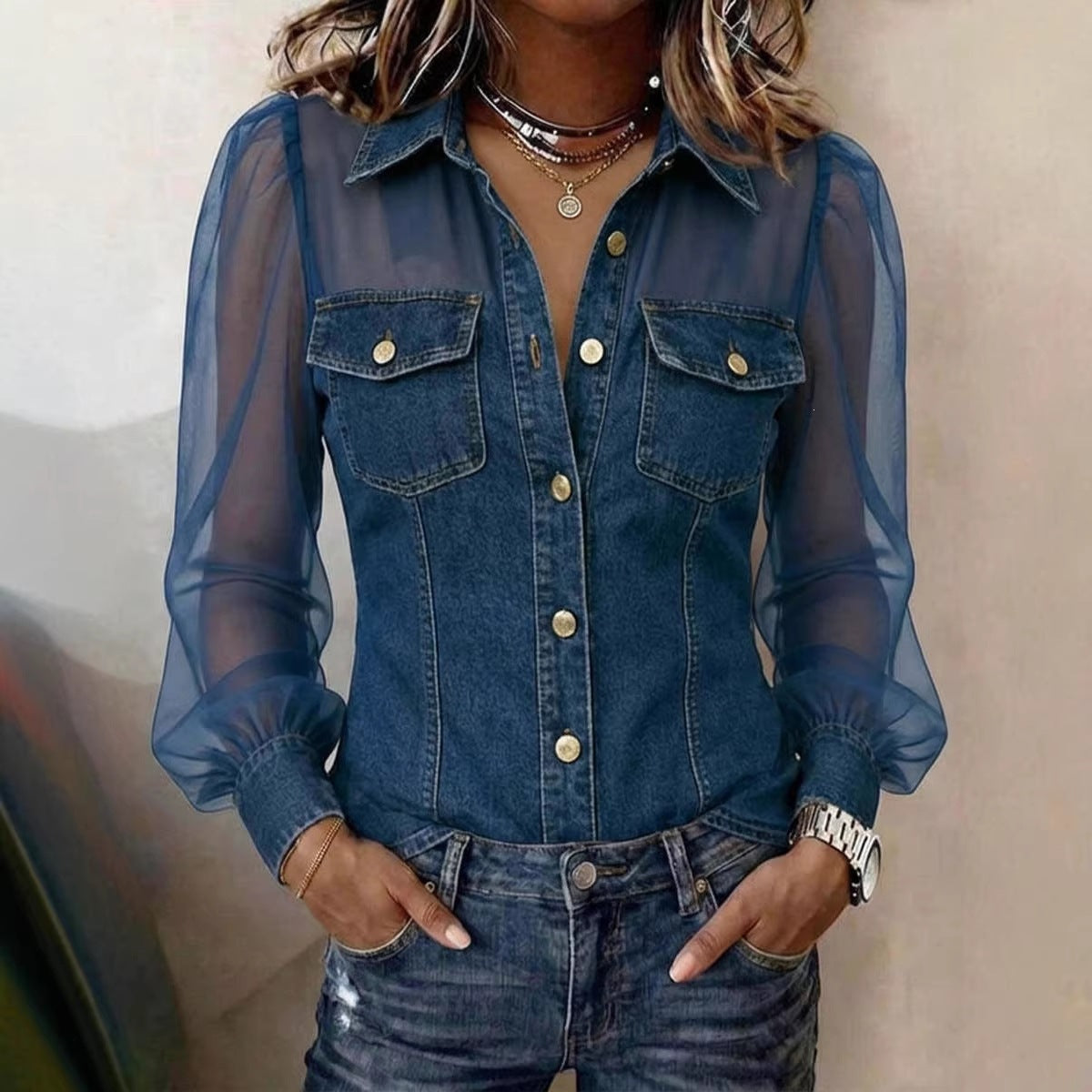 A woman wearing a blue denim long-sleeve button-up shirt with sheer mesh sleeves and gold-tone buttons, paired with dark blue jeans and layered necklaces.
