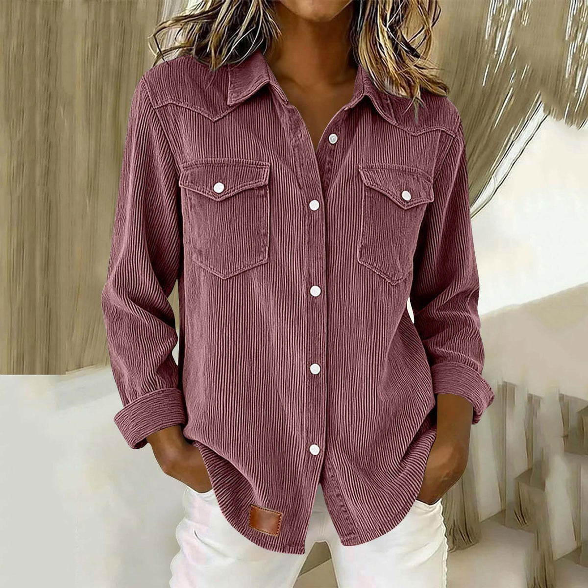 A woman wearing a long-sleeve corduroy button-up shirt in a dusty rose color, paired with white pants, shown in a styled indoor setting.