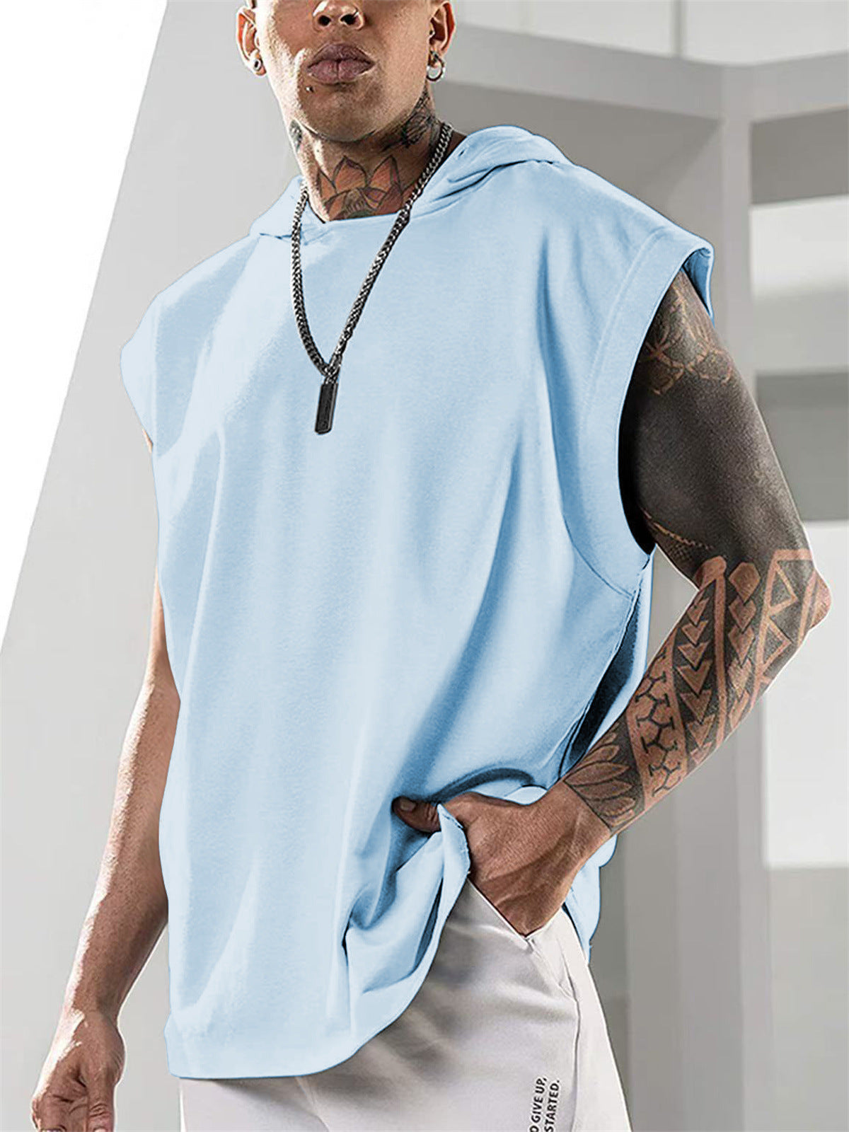 Person wearing a light blue sleeveless hoodie with tattoos on arms, standing against a neutral background.
