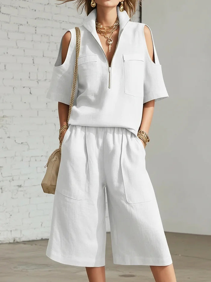 Person wearing a white outfit with cut-out shoulders against a white brick wall.