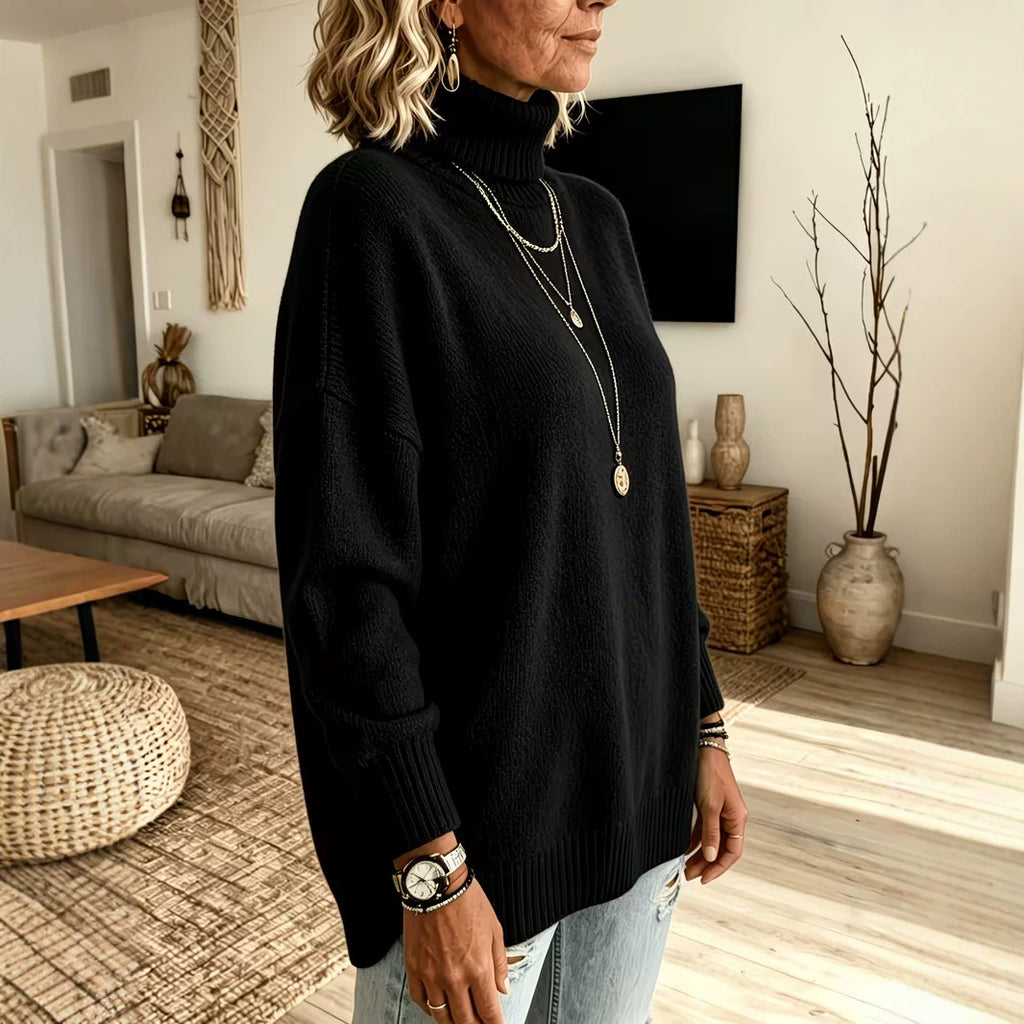 A woman wearing a black knit turtleneck sweater in a modern living room setting, paired with light-wash jeans and layered gold necklaces.