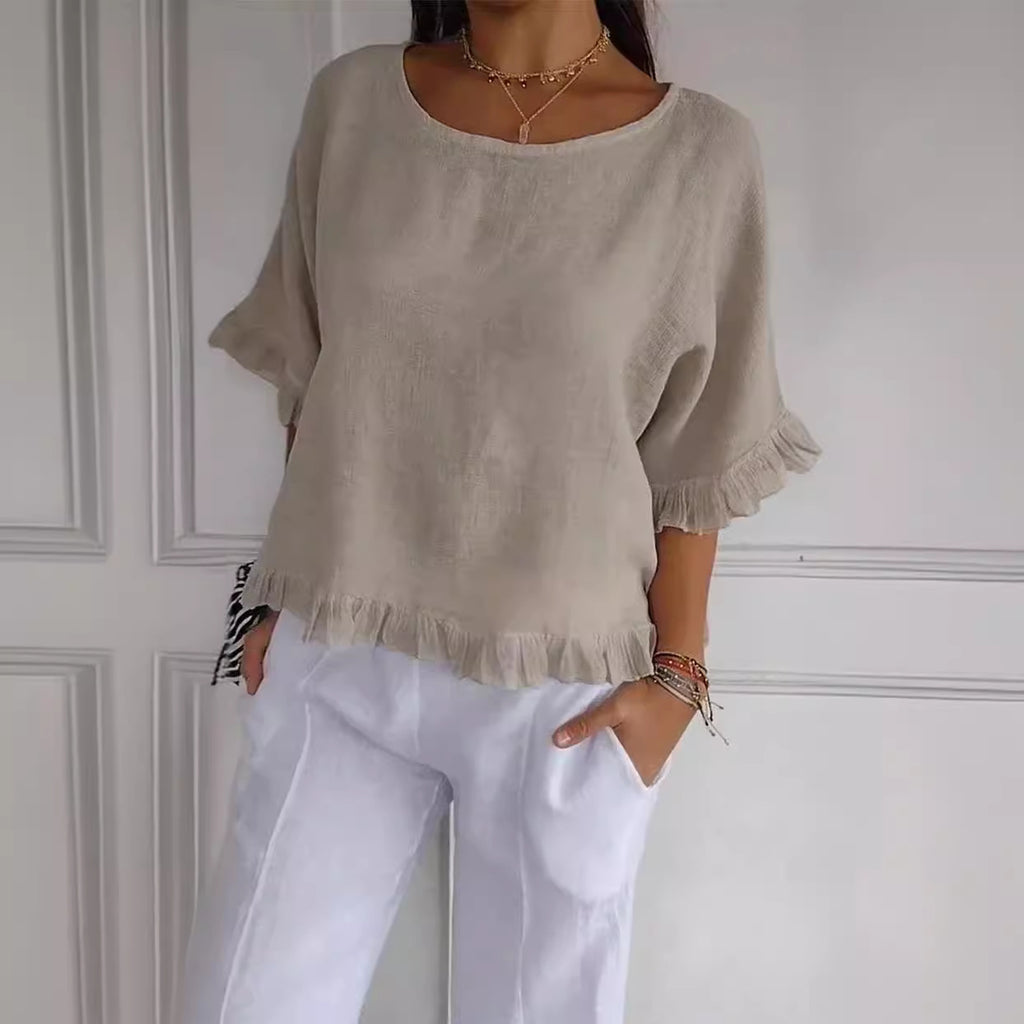 Person wearing a beige top with ruffled sleeves and white pants against a white wall.