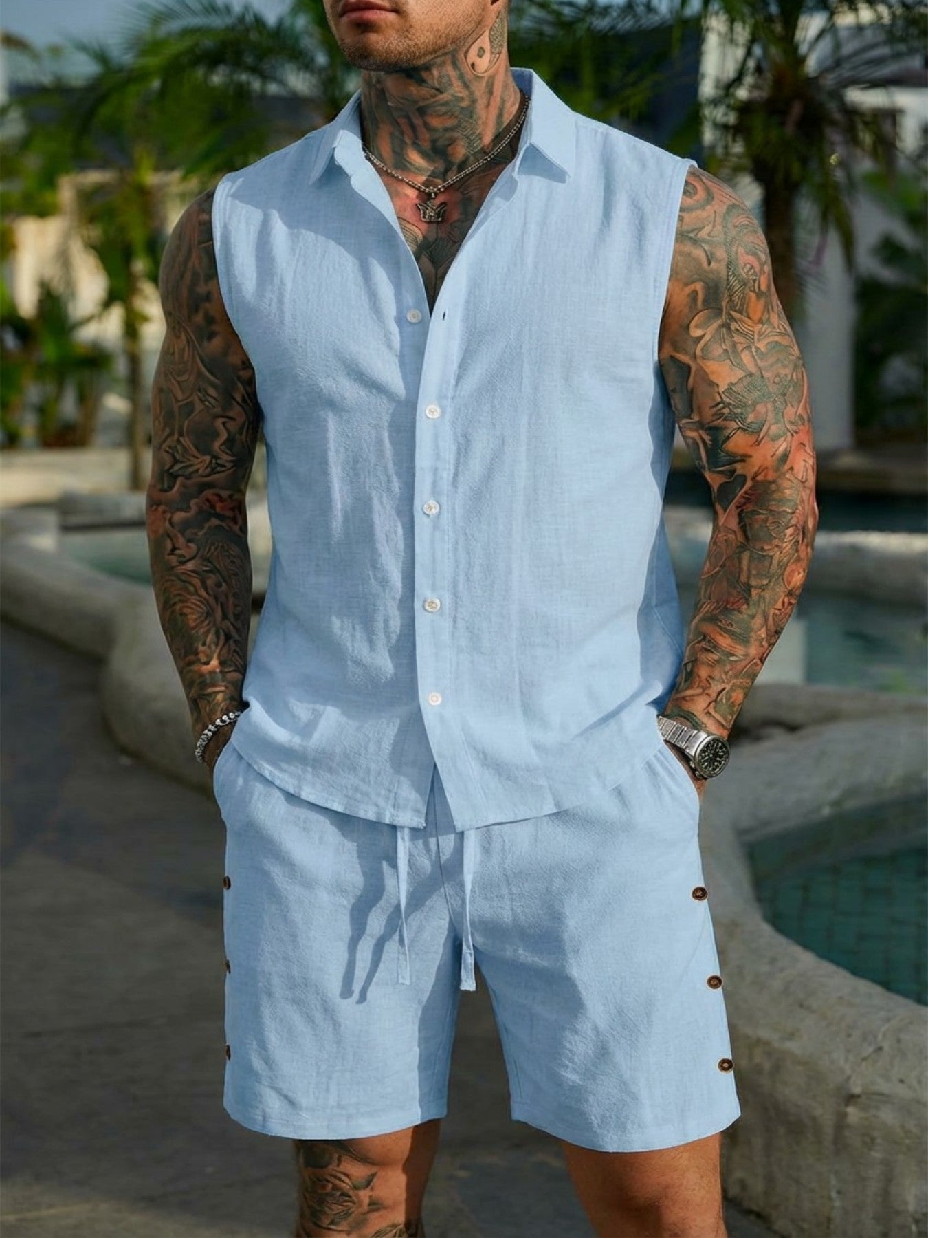 Man wearing a light blue sleeveless shirt and shorts set with tattoos on his arms, standing by a pool.