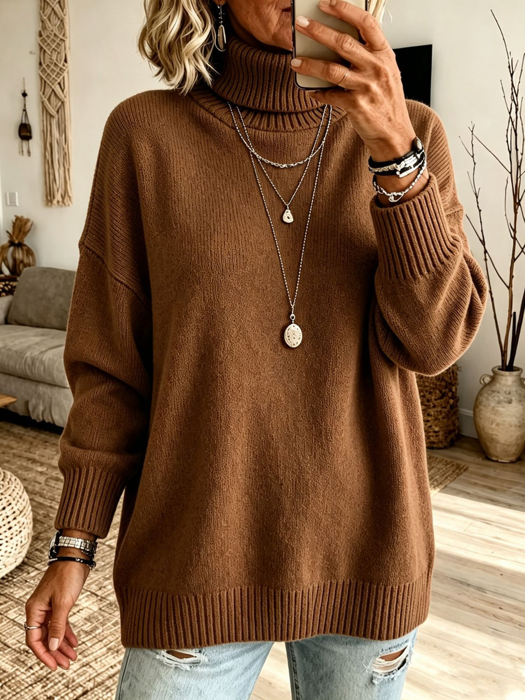 A woman wearing a brown ribbed turtleneck sweater, styled with layered silver necklaces, bracelets, and distressed light blue jeans, taking a mirror selfie in a cozy living room setting.
