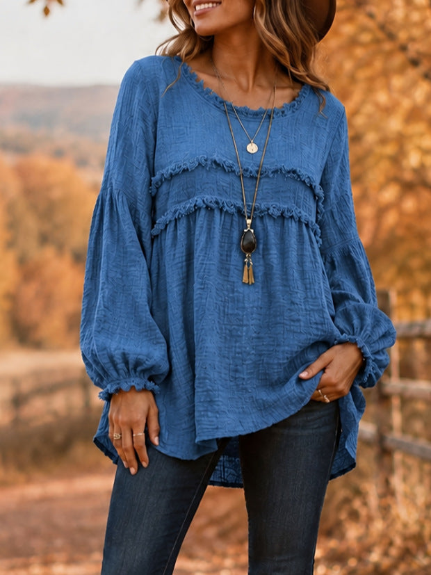 A woman wearing a blue long-sleeve blouse with layered ruffle details and elastic cuffs, paired with dark jeans, standing outdoors against a blurred autumn landscape.