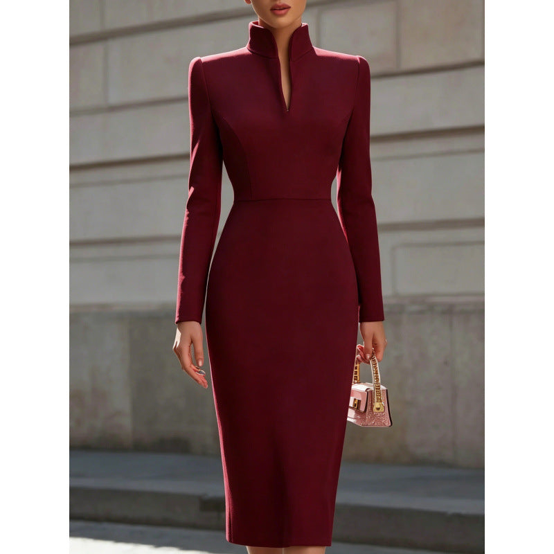 A woman wearing a fitted long-sleeve midi dress in deep burgundy with a high mock neck and structured shoulders, standing outdoors against a light stone wall and holding a small pink handbag.