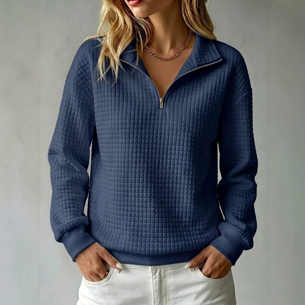 A woman wearing a navy blue waffle-knit quarter-zip pullover sweater with white pants, standing against a neutral gray wall.