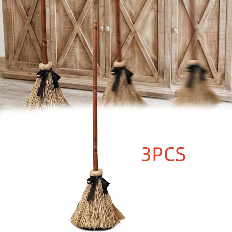 Three miniature straw brooms with brown handles and black ribbon bows, shown against a white foreground with a rustic wooden door in the background. Red text reads '3PCS'.