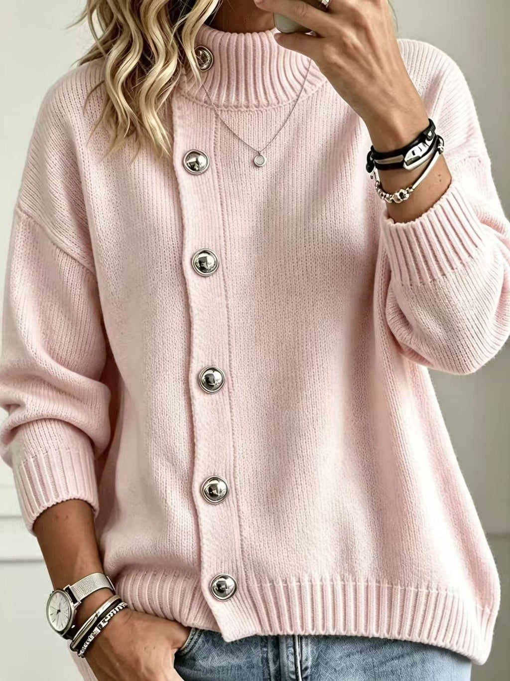 A woman wearing a light pink ribbed knit cardigan sweater with large silver buttons, paired with blue jeans and accessorized with bracelets and a necklace.
