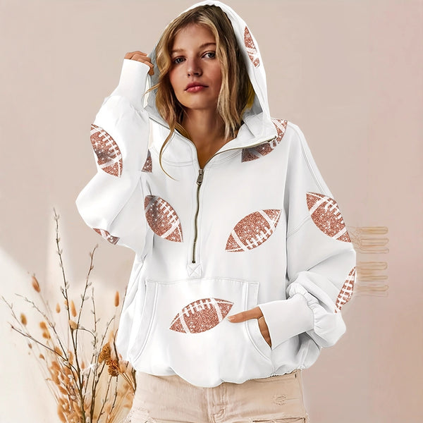 A woman wearing a white half-zip hoodie with rose gold glitter football prints, paired with beige pants, set against a soft beige background with dried grass decor.